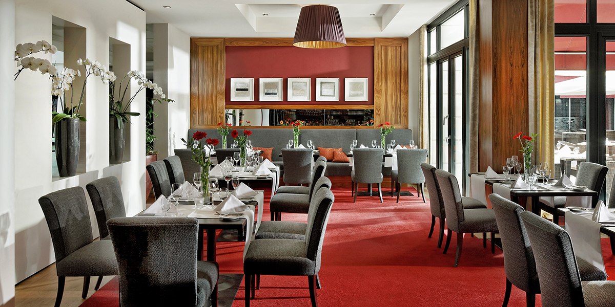 Elegantes Restaurant mit grauen Stühlen, roten Teppich und weißen Tischdecken. Große Fenster und Orchideen schmücken den Raum., © Parkhotel Stuttgart Messe-Airport