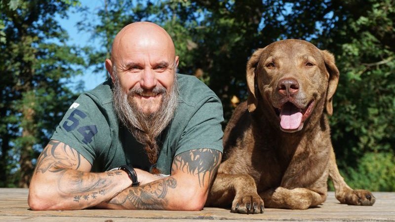 Ein Mann mit Bart und Tattoos liegt neben einem braunen Hund auf einem Holzdeck. Beide l&auml;cheln in die Kamera, umgeben von gr&uuml;nen B&auml;umen., &copy; Stadthalle Leonberg