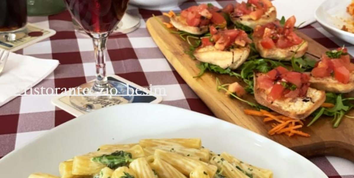 Ein Tisch mit Rigatoni in Sahnesoße, Bruschetta, Salat und Getränken auf rot-weiß karierter Tischdecke im Ristorante Zio Besim.