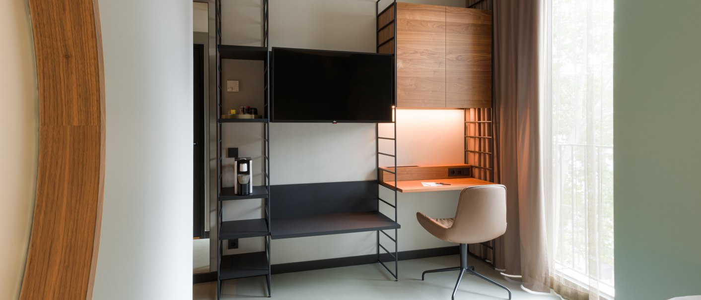 Modernes Zimmer mit Schreibtisch, Stuhl, Fernseher und gro&szlig;em Fenster. Helle, minimalistische Einrichtung mit Holz- und Metallelementen., &copy; Emilu Hotel GmbH