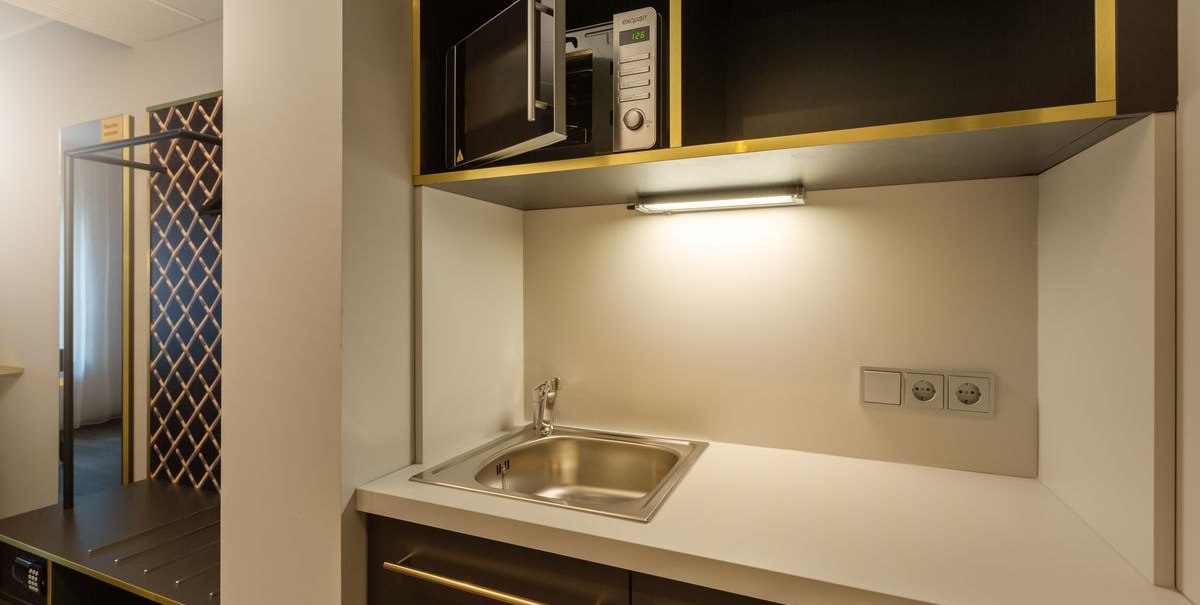 Kompakte Küche mit Spüle, Mikrowelle und Steckdosen. Dunkle Schränke mit goldenen Akzenten und Beleuchtung über der Arbeitsfläche., © Novum Hospitality Kompakte Küche mit Spüle, Mikrowelle und Steckdosen. Dunkle Schränke mit goldenen Akzenten und Beleuchtung über der Arbeitsfläche., © Novum Hospitality