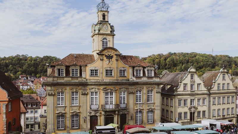 Historisches Gebäude in Schwäbisch Hall mit Marktständen davor, umgeben von weiteren alten Gebäuden und Bäumen im Hintergrund., © Nico Kurth