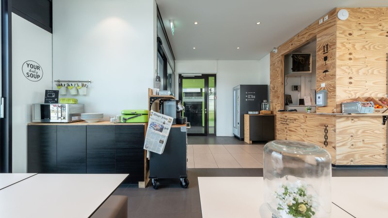 Moderne Hotel-Lounge mit Holzwänden, Snackbereich und Tisch mit Glasglocke. Zeitung und Kaffeemaschine sichtbar. Helle, einladende Atmosphäre., © a2 HOTELS Moderne Hotel-Lounge mit Holzwänden, Snackbereich und Tisch mit Glasglocke. Zeitung und Kaffeemaschine sichtbar. Helle, einladende Atmosphäre., © a2 HOTELS