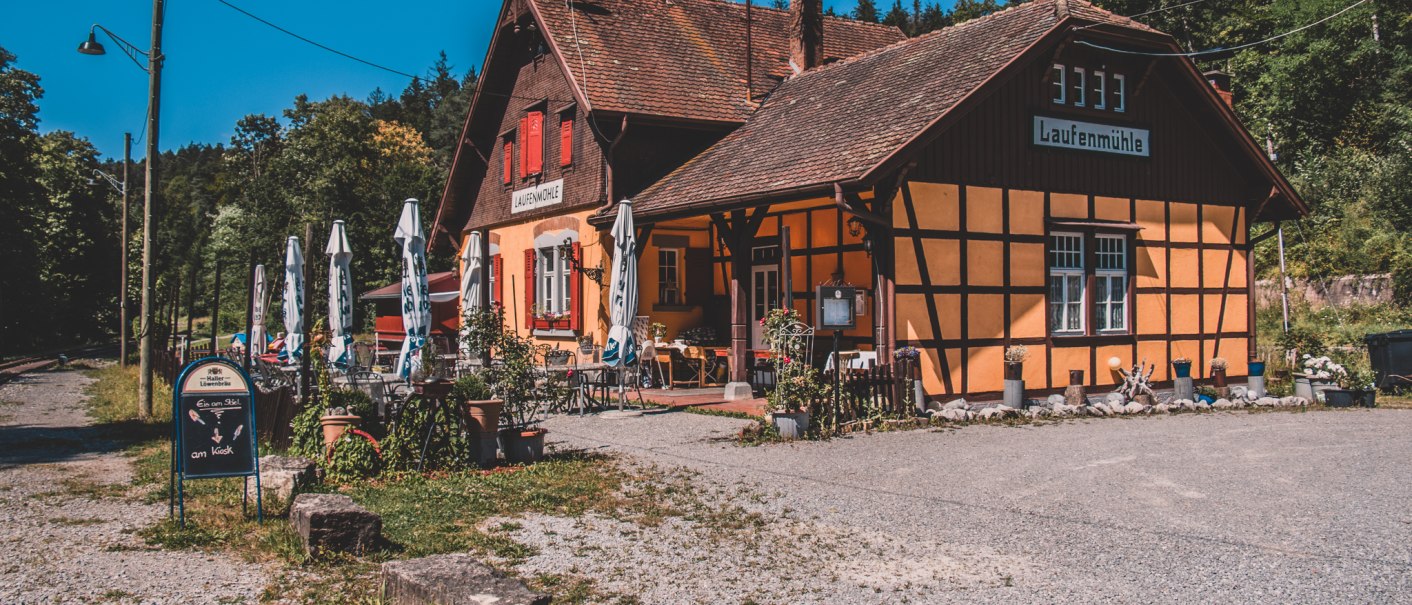 Fachwerkgebäude 'Laufenmühle' mit Sonnenschirmen und Außenbereich, umgeben von Wald, unter blauem Himmel., © Stadt Welzheim Fachwerkgebäude 'Laufenmühle' mit Sonnenschirmen und Außenbereich, umgeben von Wald, unter blauem Himmel., © Stadt Welzheim