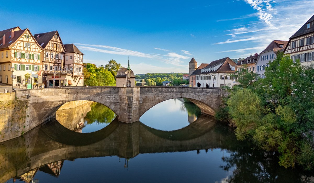 Die Henkersbrücke in Schwäbisch Hall, umgeben von Fachwerkhäusern, spiegelt sich im ruhigen Wasser unter einem klaren blauen Himmel., © Michael Kühneisen Die Henkersbrücke in Schwäbisch Hall, umgeben von Fachwerkhäusern, spiegelt sich im ruhigen Wasser unter einem klaren blauen Himmel., © Michael Kühneisen