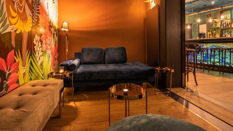 Gemütliche Lounge mit blauer Couch, buntem Wandbild mit Affen und Pflanzen. Im Hintergrund eine Bar mit beleuchteten Regalen und Hockern., © Holzmaler, Stuttgart Gemütliche Lounge mit blauer Couch, buntem Wandbild mit Affen und Pflanzen. Im Hintergrund eine Bar mit beleuchteten Regalen und Hockern., © Holzmaler, Stuttgart