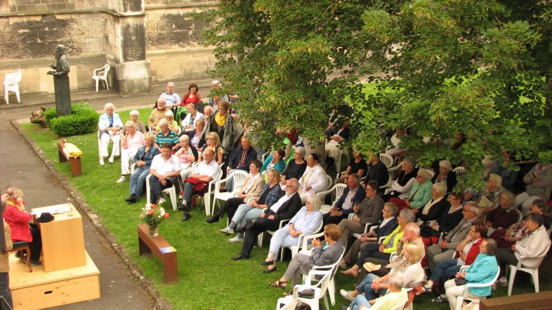 Menschen sitzen im Freien auf wei&szlig;en St&uuml;hlen und h&ouml;ren einer Lesung zu. Die Veranstaltung findet in einem historischen Hof statt, umgeben von B&auml;umen., &copy; Kulturreferat Bad Urach