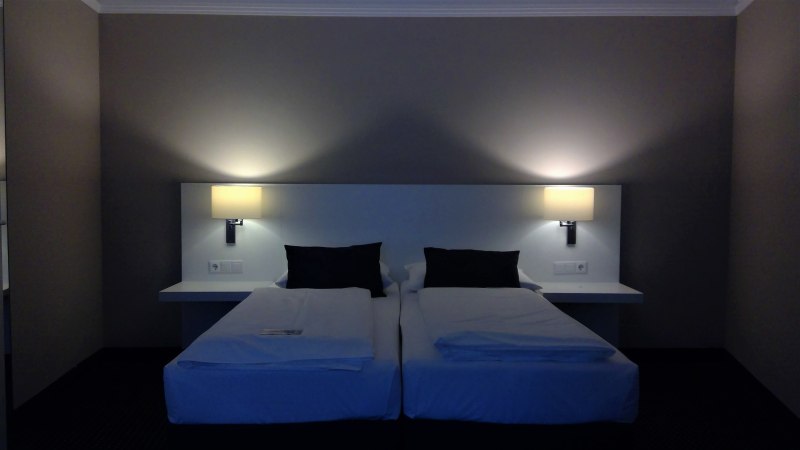 Zwei Einzelbetten mit weißen Laken und schwarzen Kissen in einem Hotelzimmer. An der Wand sind zwei Lampen angebracht, die warmes Licht spenden., © relexa Waldhotel Schatten Zwei Einzelbetten mit weißen Laken und schwarzen Kissen in einem Hotelzimmer. An der Wand sind zwei Lampen angebracht, die warmes Licht spenden., © relexa Waldhotel Schatten