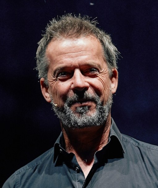 Ein Mann mit grauem Bart und Mikrofon lächelt vor einem dunklen Hintergrund. Er trägt ein dunkles Hemd., © Stadtverwaltung Remseck am Neckar