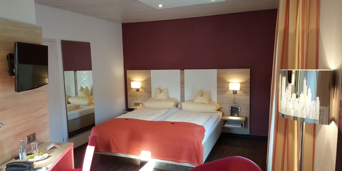 Modernes Doppelzimmer mit zwei Betten, roter Wand, Fernseher und stilvoller Beleuchtung. Ein Tisch mit Wasserflasche und Gläsern steht bereit., © Hotel Koch