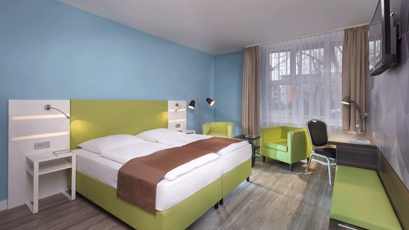 Modernes Doppelzimmer mit grünem Bett, Schreibtisch, zwei Sesseln und TV. Helle Wände und große Fenster sorgen für eine freundliche Atmosphäre., © TOMAS Modernes Doppelzimmer mit grünem Bett, Schreibtisch, zwei Sesseln und TV. Helle Wände und große Fenster sorgen für eine freundliche Atmosphäre., © TOMAS