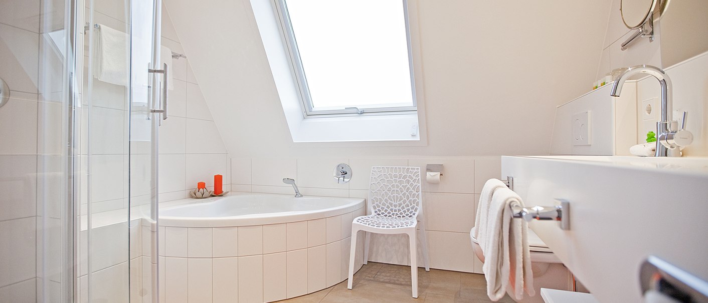 Helles Badezimmer mit Eckbadewanne, Glasdusche, Waschbecken und Dachfenster. Ein weißer Stuhl und ein orangefarbener Teppich ergänzen das Design., © Hotel Krone Helles Badezimmer mit Eckbadewanne, Glasdusche, Waschbecken und Dachfenster. Ein weißer Stuhl und ein orangefarbener Teppich ergänzen das Design., © Hotel Krone