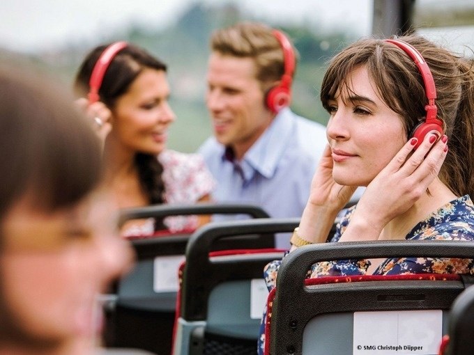 Personen in einem offenen Tourbus tragen rote Kopfh&ouml;rer und h&ouml;ren einem Audioguide zu. Im Hintergrund sind verschwommene Landschaften zu sehen., &copy; SMG, Christoph D&uuml;pper