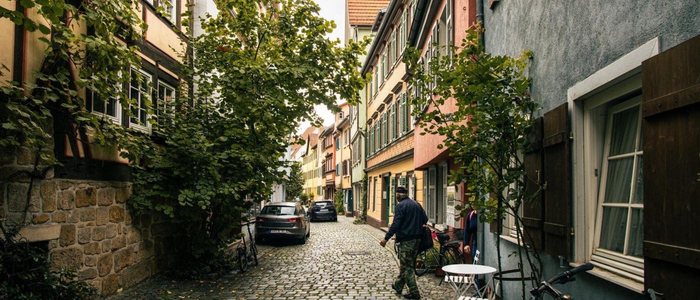 Eine idyllische Gasse in Esslingen mit Kopfsteinpflaster, Fachwerkhäusern und üppigem Grün. Autos parken am Straßenrand, während Passanten vorbeigehen., © Stuttgart-Marketing GmbH, Sarah Schmid Eine idyllische Gasse in Esslingen mit Kopfsteinpflaster, Fachwerkhäusern und üppigem Grün. Autos parken am Straßenrand, während Passanten vorbeigehen., © Stuttgart-Marketing GmbH, Sarah Schmid