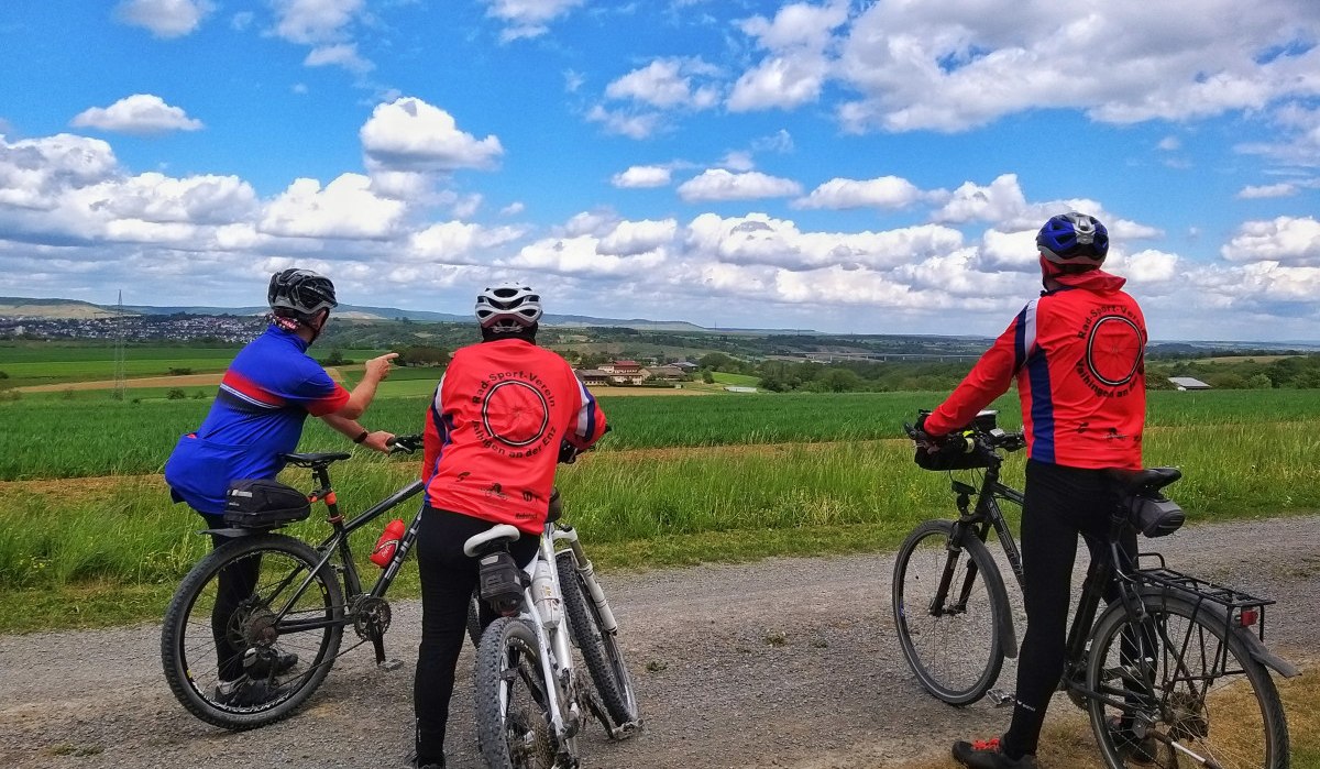 Drei Radfahrer in bunter Sportkleidung betrachten eine weite Landschaft mit Feldern und Hügeln unter einem bewölkten Himmel., © Land der 1000 Hügel - Kraichgau-Stromberg Drei Radfahrer in bunter Sportkleidung betrachten eine weite Landschaft mit Feldern und Hügeln unter einem bewölkten Himmel., © Land der 1000 Hügel - Kraichgau-Stromberg