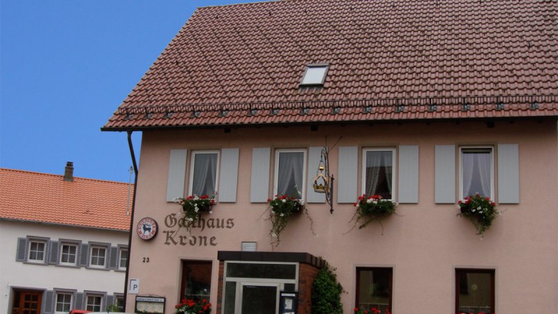 Gasthaus Krone mit blumengeschm&uuml;ckten Fenstern, zwei geparkte Autos davor, blauer Himmel im Hintergrund.