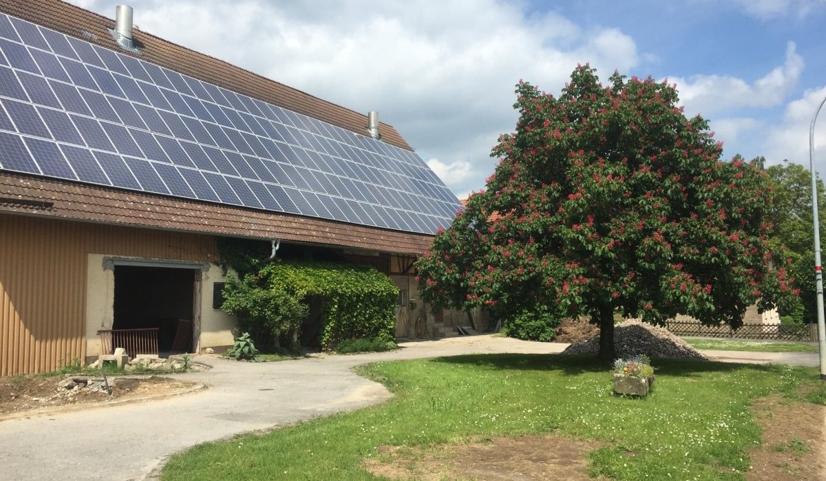 Ein Bauernhof mit Solarpanelen auf dem Dach. Ein bl&uuml;hender Baum steht im Vordergrund auf einer gr&uuml;nen Wiese unter blauem Himmel., &copy; www.pro-cycl.de