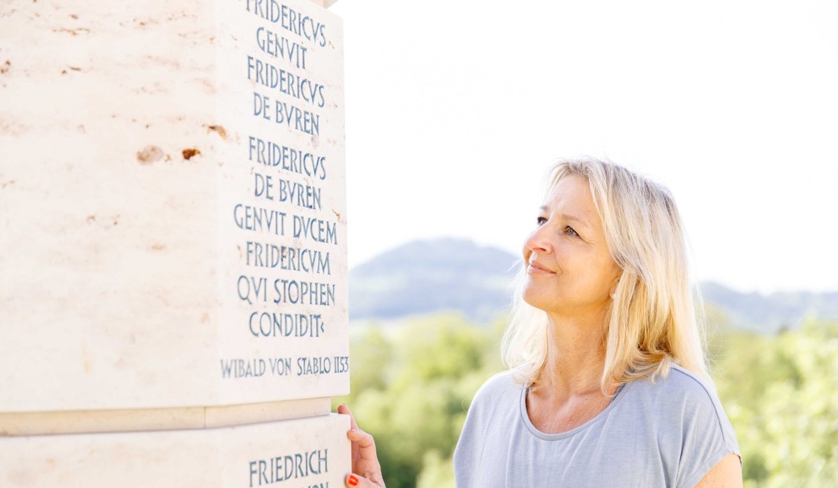 Eine Frau betrachtet eine Stele mit lateinischer Inschrift. Im Hintergrund sind Bäume und Hügel zu sehen., © Landkreis Göppingen Eine Frau betrachtet eine Stele mit lateinischer Inschrift. Im Hintergrund sind Bäume und Hügel zu sehen., © Landkreis Göppingen
