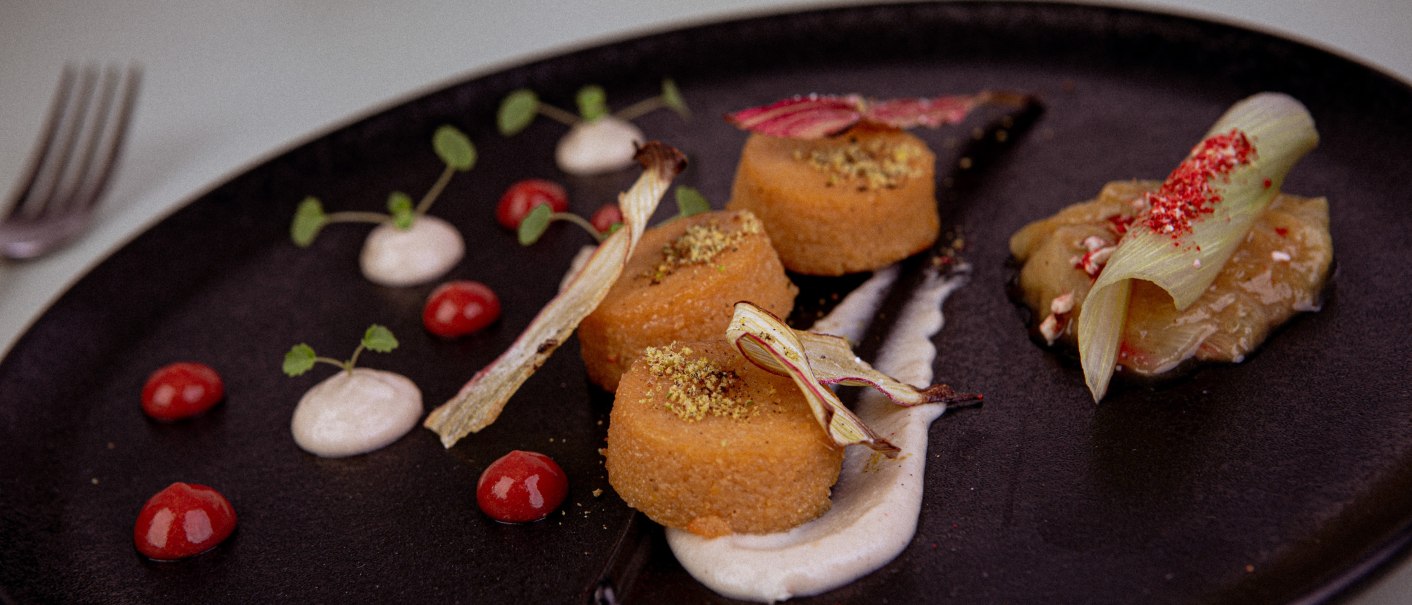 Gourmetgericht auf schwarzem Teller, dekoriert mit Saucen, Kräutern und Gemüse. Elegante Anordnung mit verschiedenen Texturen und Farben., © Marc Luppa