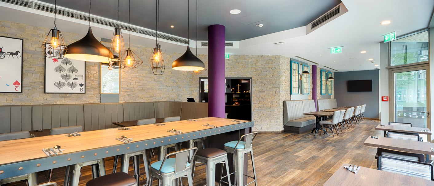 Moderner Frühstücksbereich mit langen Holztischen, Metallstühlen und stilvollen Hängelampen. Steinwände und große Fenster schaffen eine einladende Atmosphäre., © Premier Inn Germany