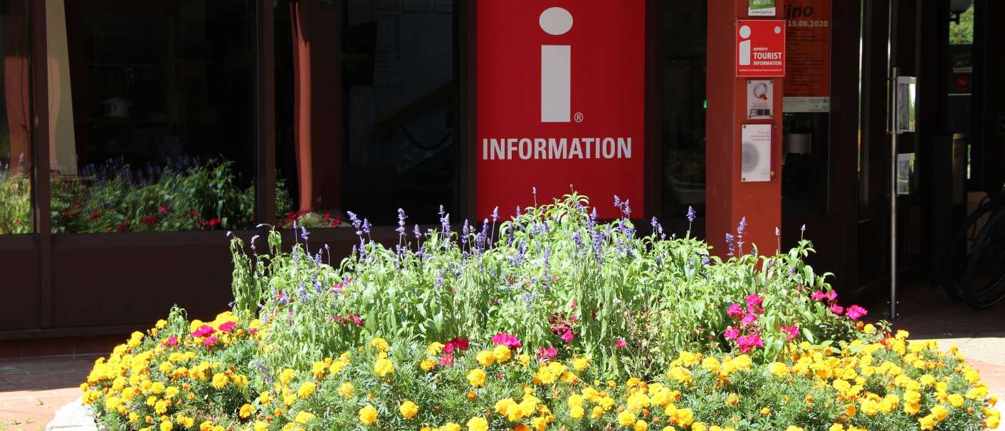 Eingang zur Tourist Information im Kurpark, umgeben von einem bunten Blumenbeet mit gelben und lila Blüten., © Bad Urach Tourismus