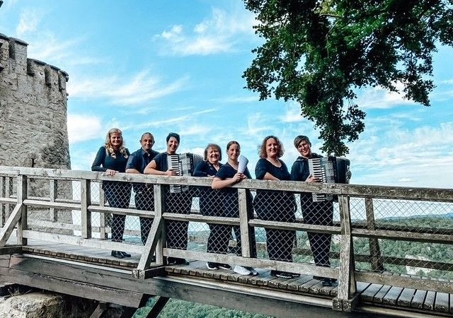 Sechs Personen mit Akkordeons stehen auf einer Holzbrücke vor einer Burgmauer, umgeben von Bäumen und blauem Himmel., © ConPassione Sechs Personen mit Akkordeons stehen auf einer Holzbrücke vor einer Burgmauer, umgeben von Bäumen und blauem Himmel., © ConPassione