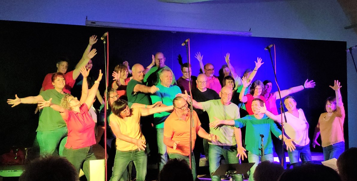 Ein Chor in bunten Outfits singt und tanzt auf einer B&uuml;hne mit lebendiger Beleuchtung.