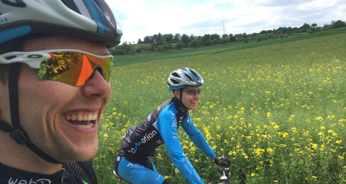 Zwei Fahrradfahrer in Sportkleidung und Helmen fahren lachend durch ein blühendes Feld unter einem bewölkten Himmel., © www.pro-cycl.de Zwei Fahrradfahrer in Sportkleidung und Helmen fahren lachend durch ein blühendes Feld unter einem bewölkten Himmel., © www.pro-cycl.de