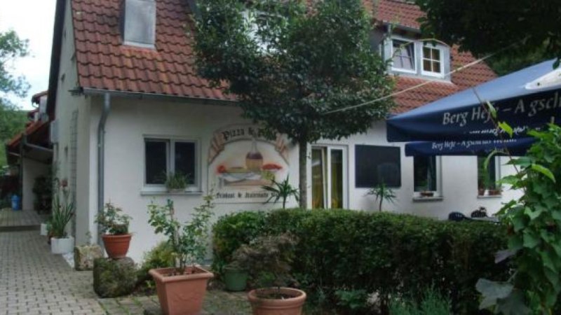 Ein gem&uuml;tliches Restaurant mit roten Dachziegeln, umgeben von Pflanzen. Ein blauer Sonnenschirm steht im Vordergrund.