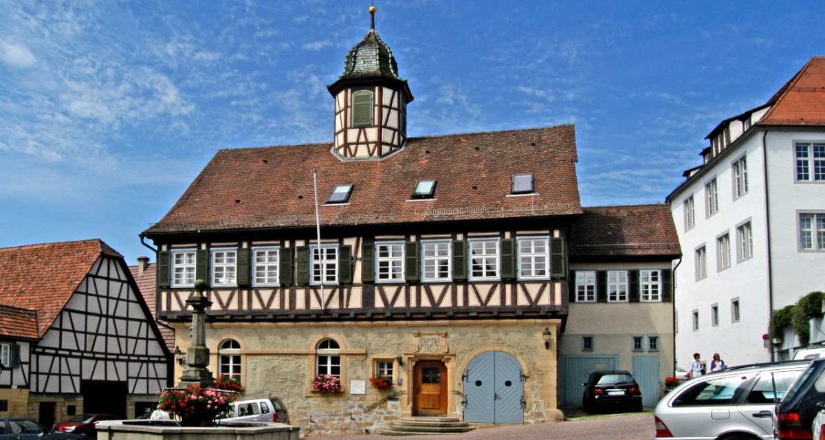 Historisches Fachwerkgeb&auml;ude mit kleinem Turm und Brunnen im Vordergrund. Autos parken auf dem Platz, blauer Himmel im Hintergrund., &copy; Natur.Nah. Sch&ouml;nbuch & Heckeng&auml;u