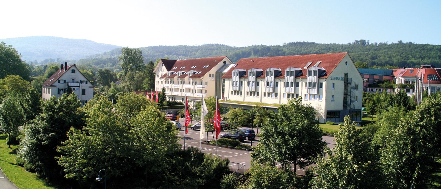 Gebäudekomplex mit roten Dächern, umgeben von Bäumen und Hügeln in Bad Boll. Mehrere Flaggen sind vor dem Gebäude zu sehen., © Seminaris Hotel Bad Boll Gebäudekomplex mit roten Dächern, umgeben von Bäumen und Hügeln in Bad Boll. Mehrere Flaggen sind vor dem Gebäude zu sehen., © Seminaris Hotel Bad Boll