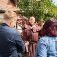 Ein M&ouml;nch in brauner Kutte f&uuml;hrt eine Gruppe von Menschen durch eine malerische Umgebung mit Fachwerkhaus und bl&uuml;henden Pflanzen., &copy; Stadtverwaltung Weil der Stadt