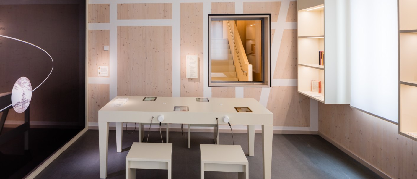 Moderne Ausstellung mit weißem Tisch und Stühlen, Regalen und einem Fenster, das eine Treppe zeigt. Die Wände sind in Holzoptik gestaltet., © (c)dieargelola