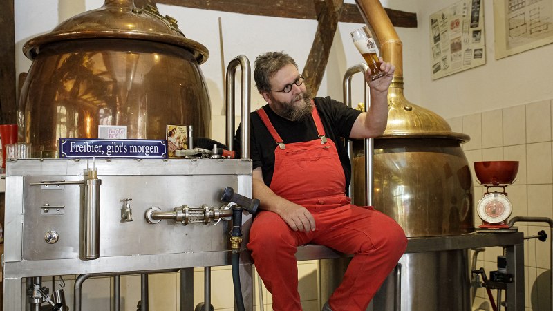 Ein Mann in roter Latzhose sitzt auf einer Kupferbrauanlage und betrachtet ein Bierglas. Im Hintergrund sind Gläser und Brauzubehör zu sehen., © Torsten Wenzler