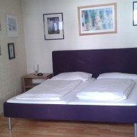 Ein gemütliches Doppelzimmer mit einem Doppelbett, umgeben von gerahmten Bildern an den Wänden. Holzboden und Nachttische sind sichtbar., © City Hotel Fellbach