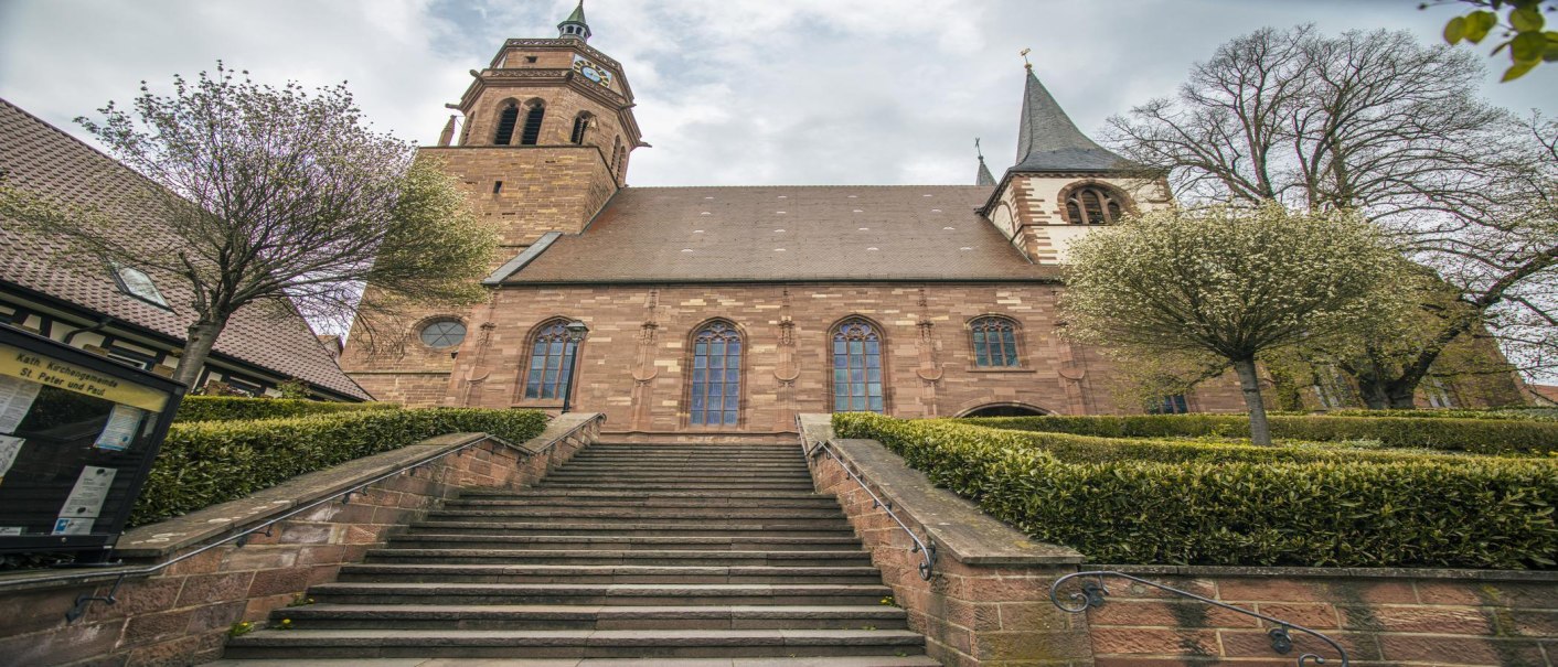 Die St. Peter und Paul Kirche in Weil der Stadt, aufgenommen im Weitwinkel. Eine Treppe führt zur Kirche, umgeben von Bäumen und Sträuchern., © SMG, Sarah Schmid