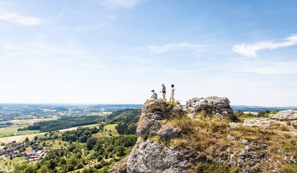 Drei Personen stehen auf einem Felsen und blicken in die weite, grüne Landschaft unter einem klaren blauen Himmel., © TMBW