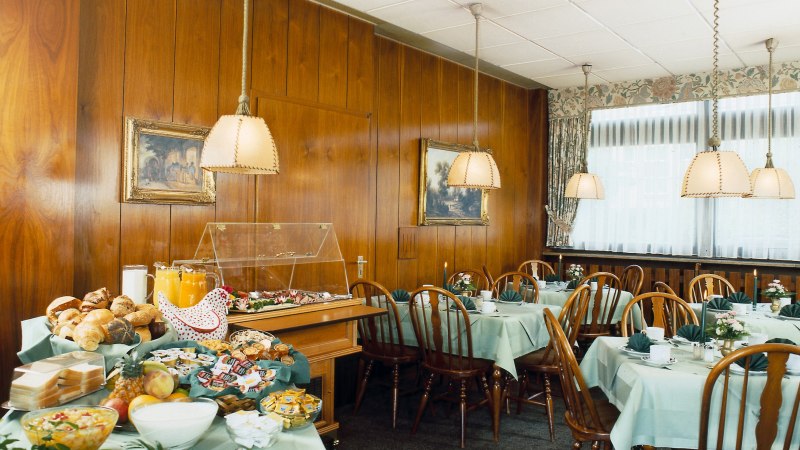Fr&uuml;hst&uuml;cksraum mit gedeckten Tischen, Holzst&uuml;hlen und einem Buffet mit Br&ouml;tchen, Obst und Saft. Holzw&auml;nde und h&auml;ngende Lampen schaffen eine gem&uuml;tliche Atmosph&auml;re., &copy; Hotel Find