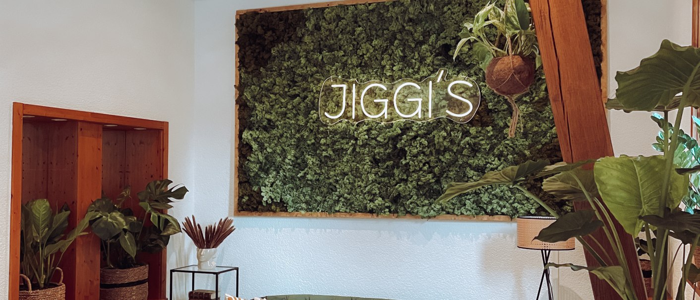 Gemütlicher Raum mit grüner Couch, Rattanmöbeln und Pflanzen. An der Wand ein Schild mit der Aufschrift 'Jiggi's'. Helle, einladende Atmosphäre., © Jiggis