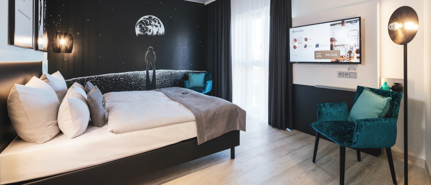 Modernes Hotelzimmer mit einem großen Bett, einem Sessel und einem Fernseher. Die Wand zeigt ein Weltraum-Motiv mit Astronaut und Mond., © Novum Management GmbH Modernes Hotelzimmer mit einem großen Bett, einem Sessel und einem Fernseher. Die Wand zeigt ein Weltraum-Motiv mit Astronaut und Mond., © Novum Management GmbH