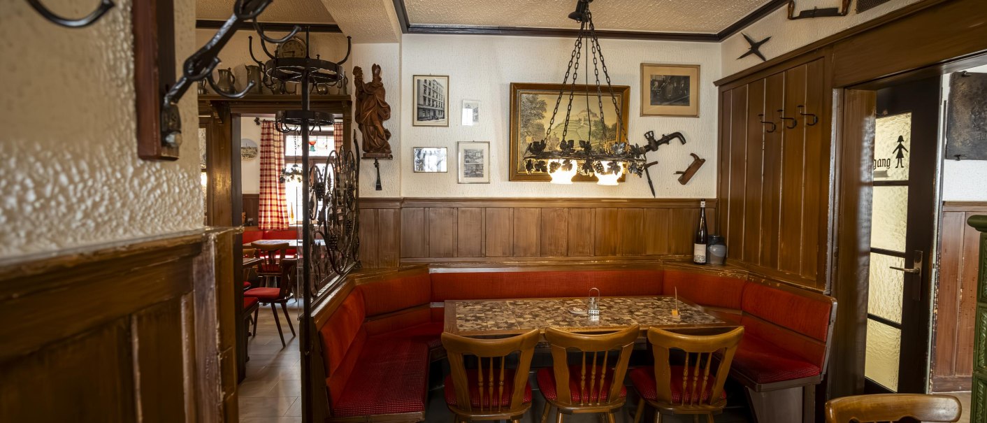 Gemütliche Weinstube mit Holzmöbeln, roten Polstern und rustikaler Dekoration. An den Wänden hängen Bilder und Werkzeuge, ein Kronleuchter beleuchtet den Raum., © Stuttgart Marketing GmbH, Sarah Schmid