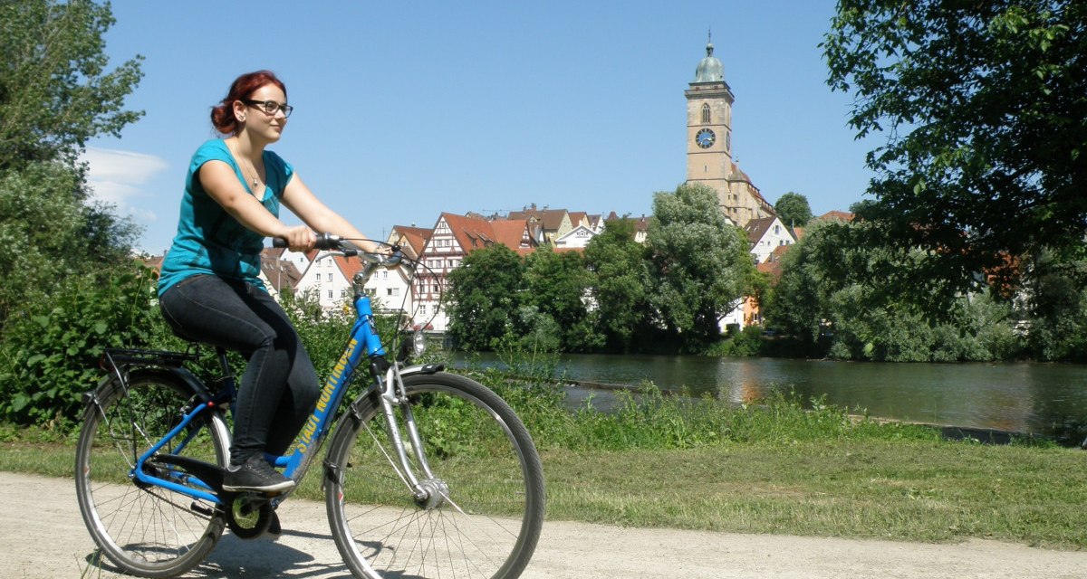 Eine Frau fährt Fahrrad am Neckar in Nürtingen. Im Hintergrund sind eine Kirche und Fachwerkhäuser zu sehen., © Stadt Nürtingen Eine Frau fährt Fahrrad am Neckar in Nürtingen. Im Hintergrund sind eine Kirche und Fachwerkhäuser zu sehen., © Stadt Nürtingen