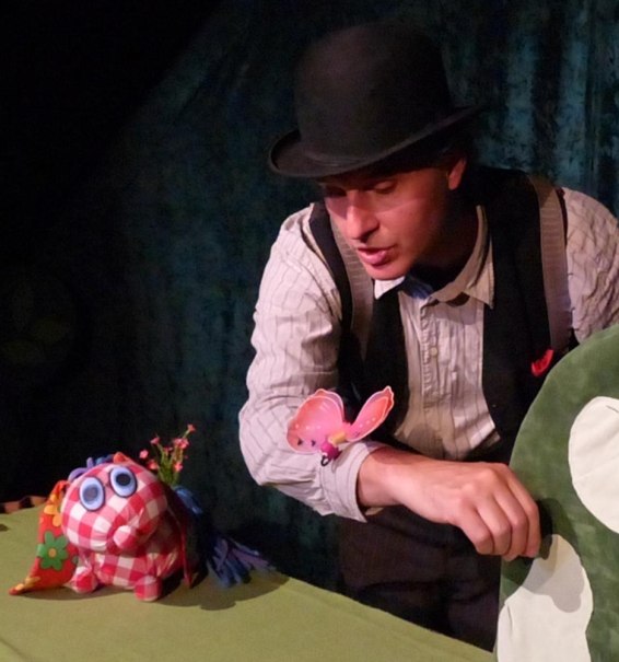 Ein Schauspieler mit Melone und Weste interagiert mit einer bunten Stofffigur und einem Schmetterling auf einer Bühne., © HalloDu-Theater