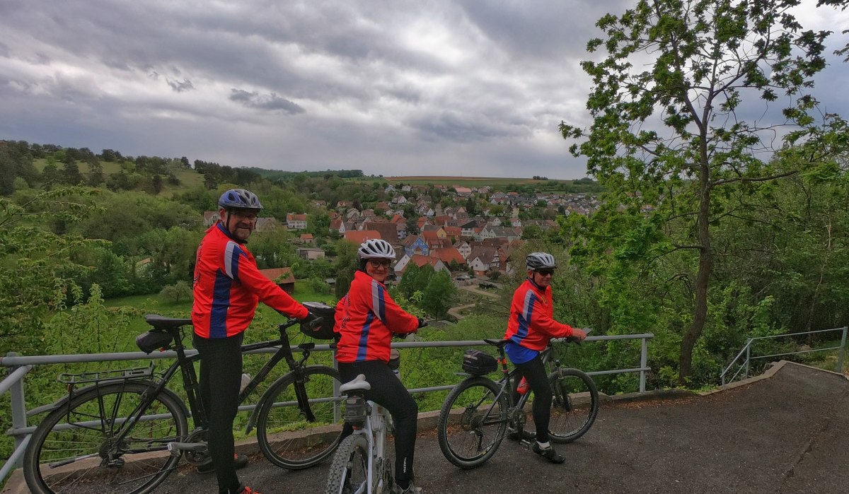 Drei Radfahrer in roten Jacken stehen auf einem Hügel und blicken auf ein Dorf im Tal. Der Himmel ist bewölkt, die Landschaft grün., © Land der 1000 Hügel - Kraichgau-Stromberg Drei Radfahrer in roten Jacken stehen auf einem Hügel und blicken auf ein Dorf im Tal. Der Himmel ist bewölkt, die Landschaft grün., © Land der 1000 Hügel - Kraichgau-Stromberg