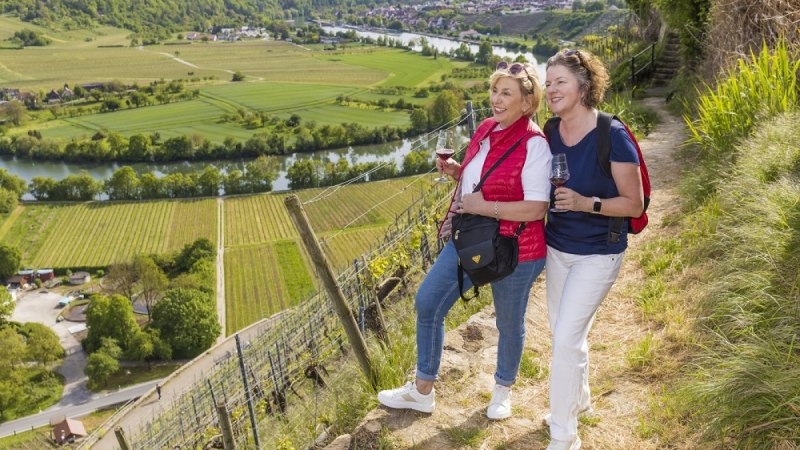 Zwei Frauen wandern mit Weingl&auml;sern durch Weinberge. Im Hintergrund sind ein Fluss und gr&uuml;ne Felder zu sehen.