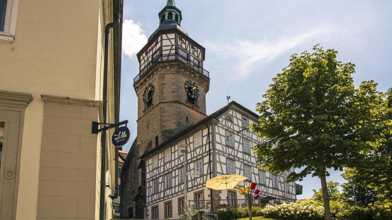 Fachwerkhaus und Kirchturm in Backnang, umgeben von Bäumen und Blumen, bei sonnigem Wetter. Ein Schild mit der Aufschrift 'Lille Bonne' ist sichtbar., © Stuttgart-Marketing GmbH, Sarah Schmid Fachwerkhaus und Kirchturm in Backnang, umgeben von Bäumen und Blumen, bei sonnigem Wetter. Ein Schild mit der Aufschrift 'Lille Bonne' ist sichtbar., © Stuttgart-Marketing GmbH, Sarah Schmid