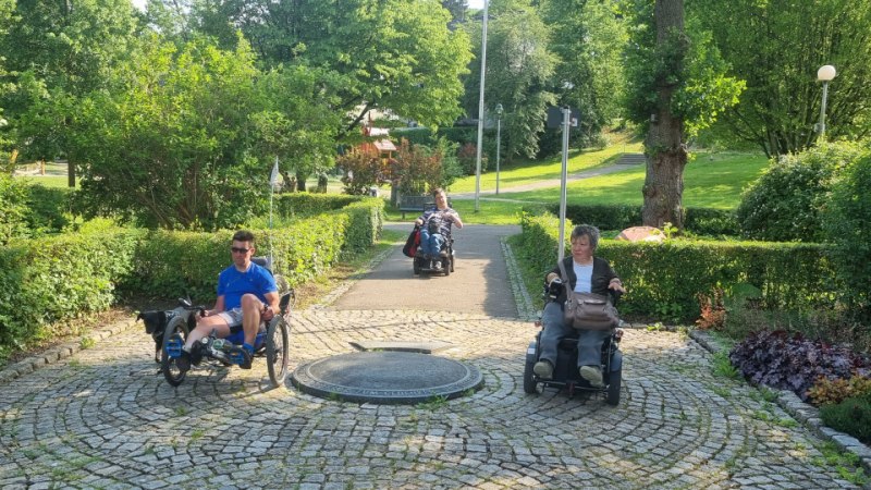 Drei Personen in Rollst&uuml;hlen fahren auf einem gepflasterten Weg durch einen gr&uuml;nen Park mit B&auml;umen und Str&auml;uchern., &copy; NPSFW