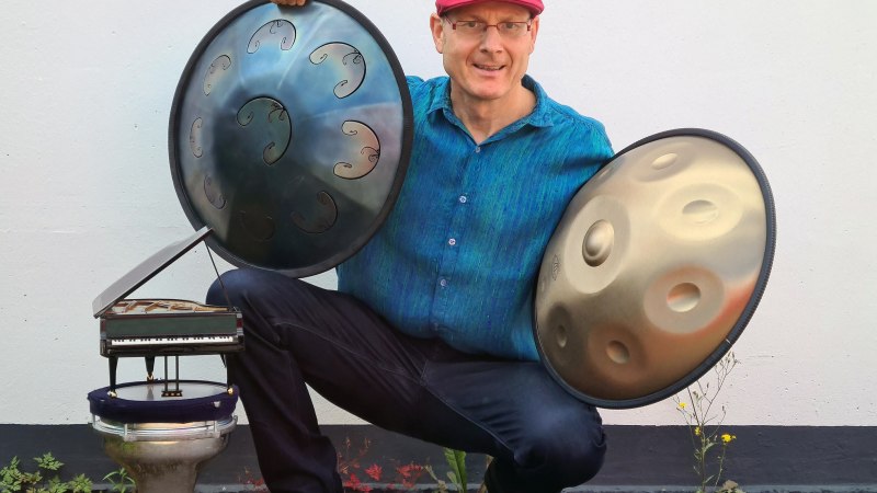 Ein Mann mit roter M&uuml;tze h&auml;lt zwei Handpans und sitzt neben einem kleinen Klaviermodell auf einer Trommel. Er tr&auml;gt ein blaues Hemd und l&auml;chelt in die Kamera., &copy; Holger Mantey