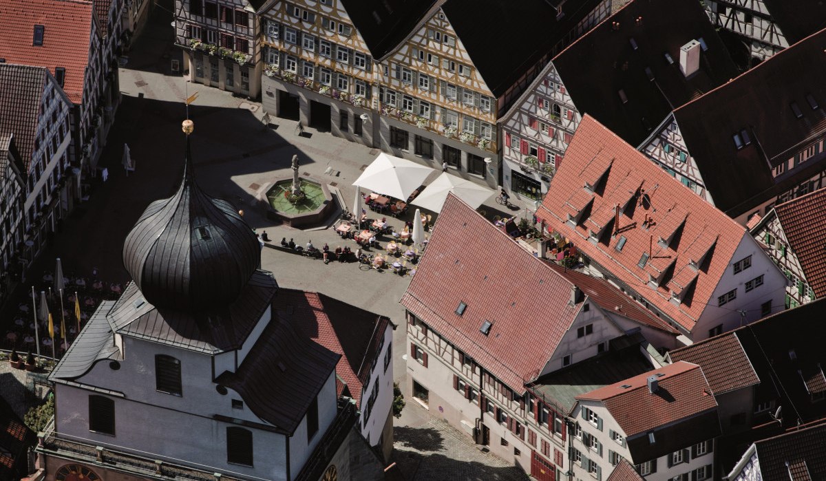 Luftaufnahme eines Marktplatzes mit Fachwerkhäusern, einem Brunnen und einem Gebäude mit Zwiebelturm. Menschen sitzen unter Sonnenschirmen., © Regio Marketing GmbH Luftaufnahme eines Marktplatzes mit Fachwerkhäusern, einem Brunnen und einem Gebäude mit Zwiebelturm. Menschen sitzen unter Sonnenschirmen., © Regio Marketing GmbH