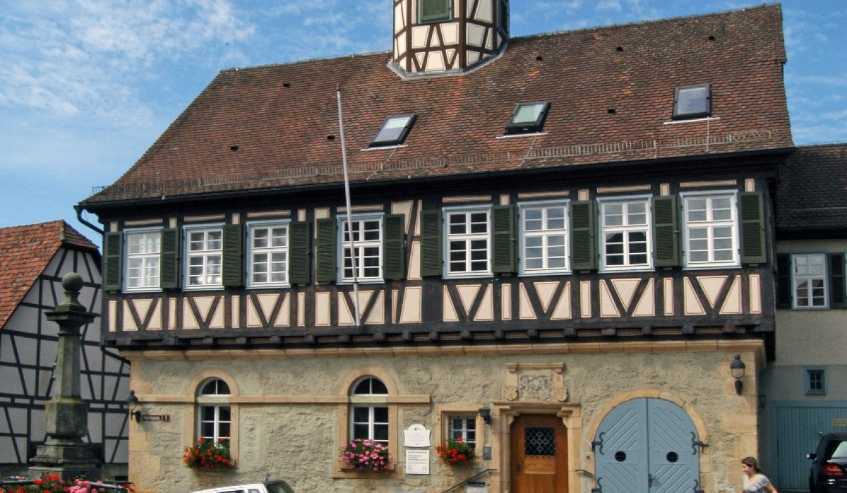 Fachwerkgebäude mit Turm und Brunnen auf gepflastertem Platz, blauer Himmel im Hintergrund., © Natur.Nah. Schönbuch & Heckengäu Fachwerkgebäude mit Turm und Brunnen auf gepflastertem Platz, blauer Himmel im Hintergrund., © Natur.Nah. Schönbuch & Heckengäu